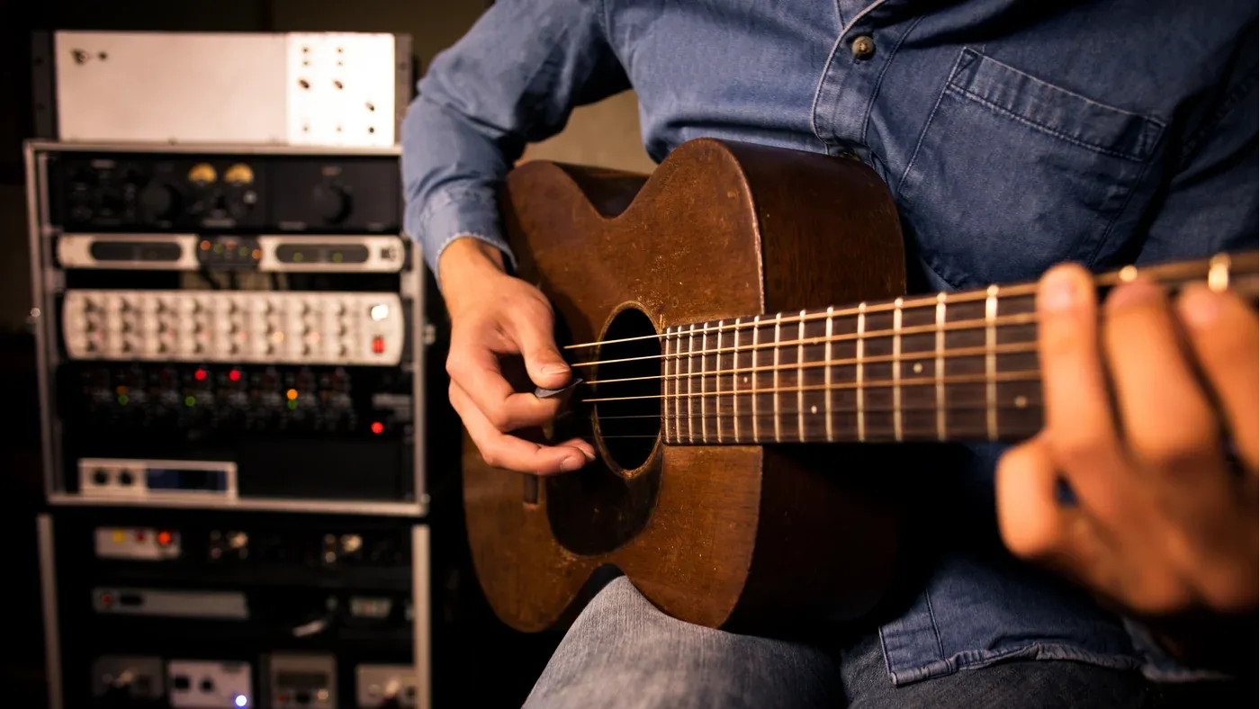 A player strumming a worn mahogany acoustic guitar, a rack of studio outboard behind them.