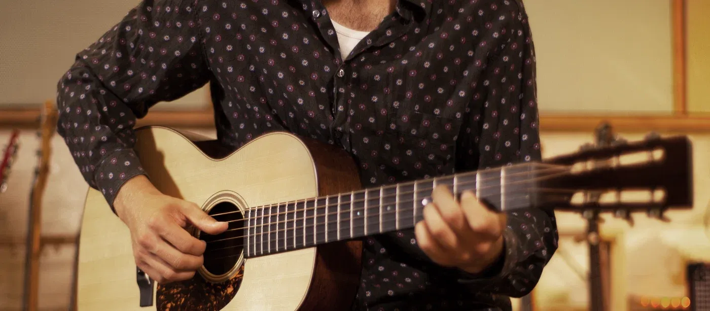 A player fingerpicking a small-body steel-string acoustic guitar in a warm studio room.