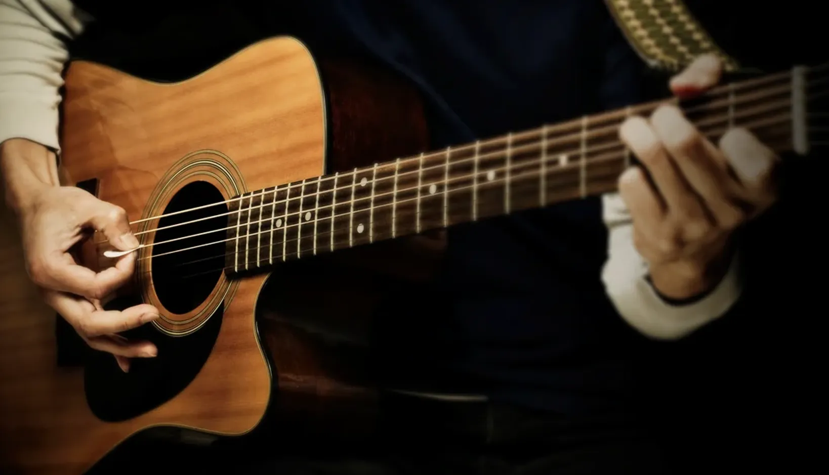 A player strumming a natural-top acoustic guitar with a pick, fretting hand high on the neck.