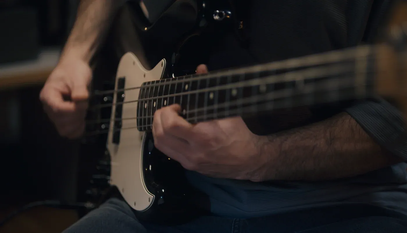 A player's hands on a black dual-pickup bass with a white scratchplate, one hand fretting and the other plucking.