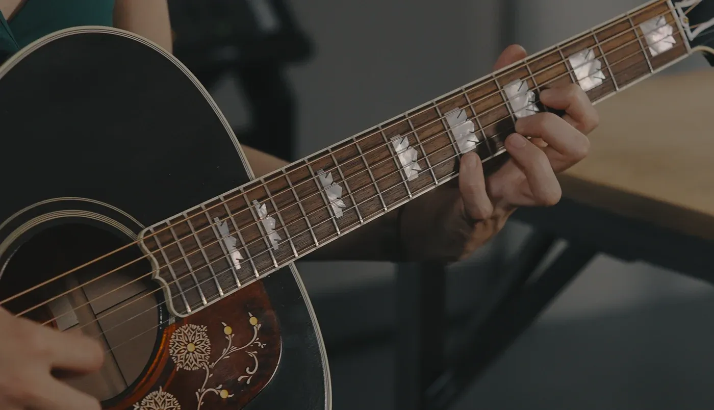 A player fretting high on the neck of a jumbo acoustic guitar with an inlaid pickguard and mother-of-pearl markers.