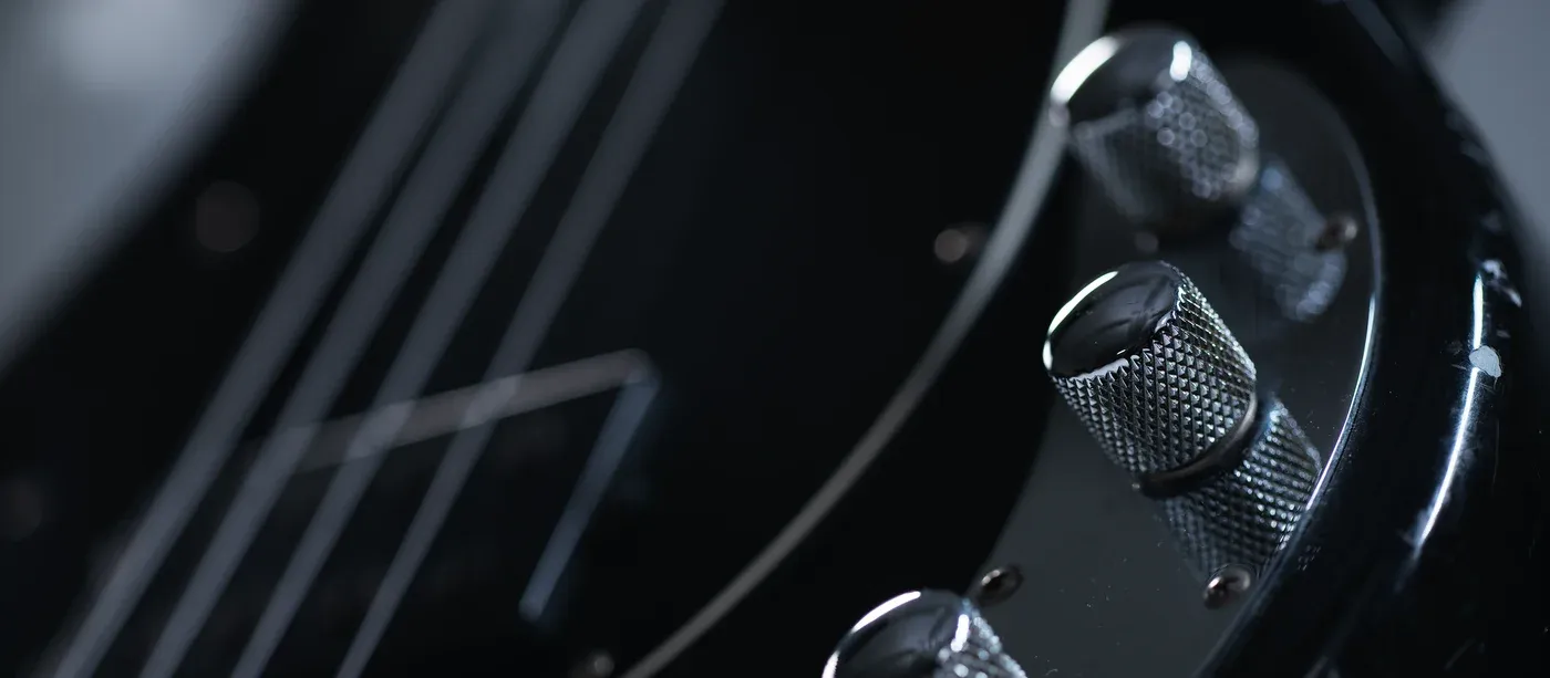A black lacquered bass photographed close along its strings, four knurled chrome control knobs catching the light.