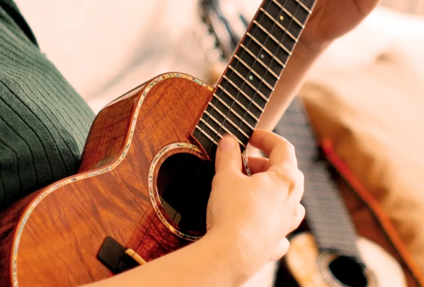 A player picking a koa ukulele with abalone binding, a second instrument resting beside them.
