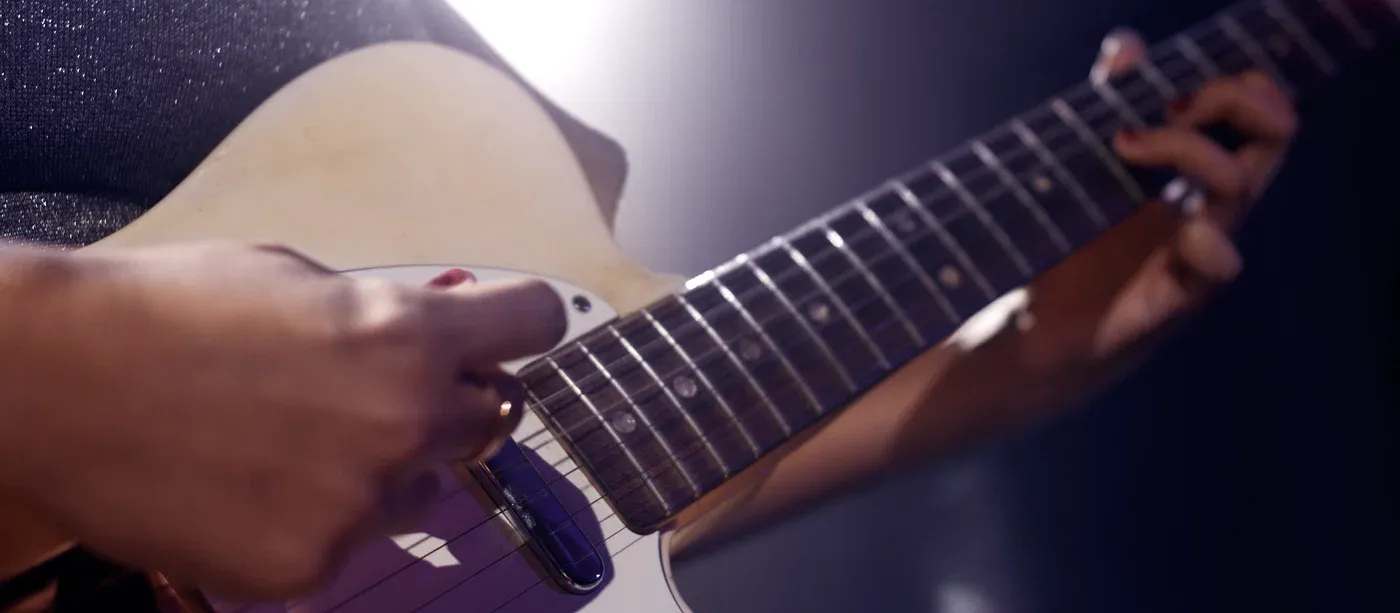 A player's hands on a blonde solid-body electric guitar under violet stage light, one hand picking and the other high up the neck.