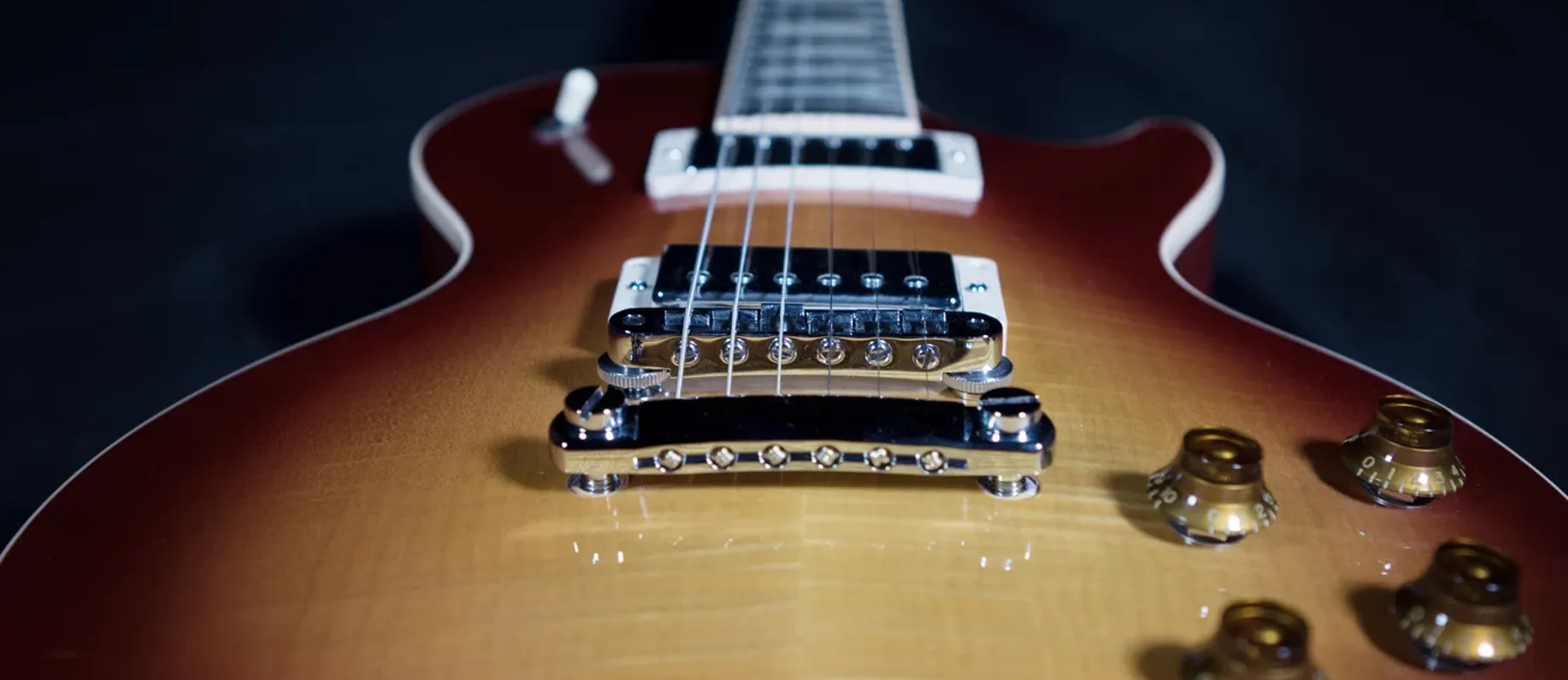 Close-up along the body of a cherry sunburst electric guitar, the tailpiece, bridge and humbucker in focus with the gold control knobs behind.