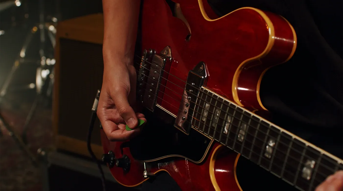 A player with a green plectrum on a cherry red hollowbody electric guitar, an amplifier behind them.