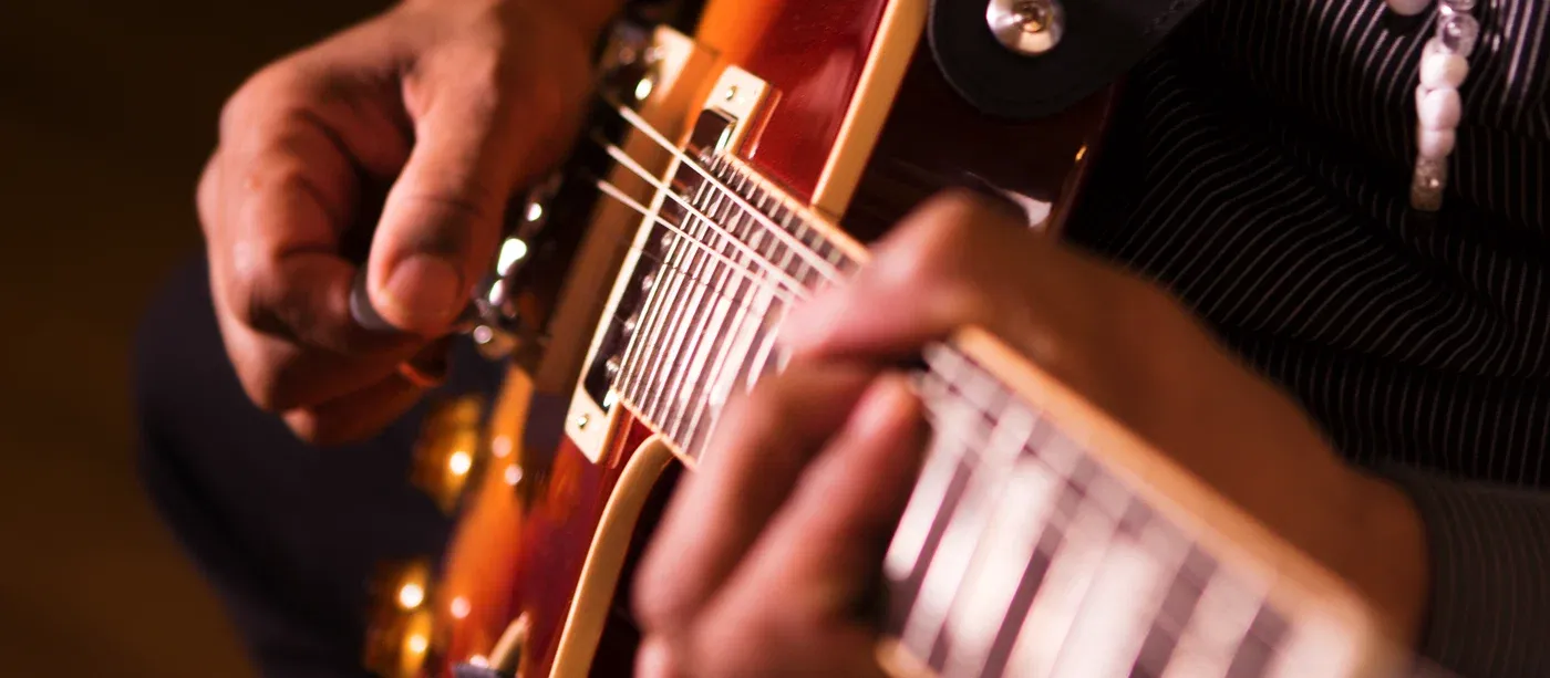 A player's hands on a cherry sunburst electric guitar, the right hand at the bridge pickup and the left hand fretting up the neck.