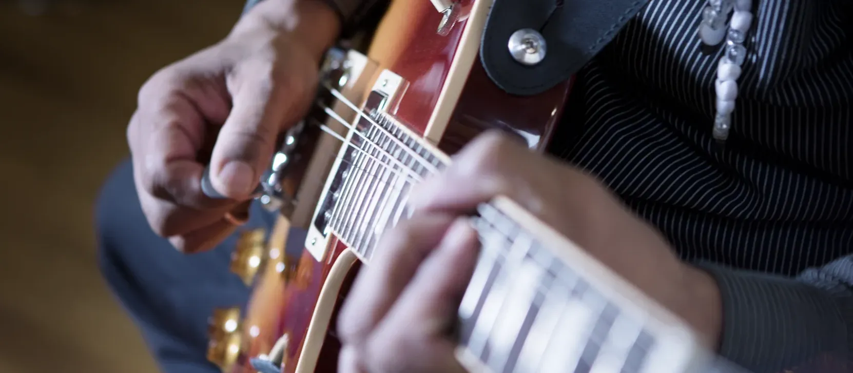 Both of a player's hands at work on a cherry sunburst electric guitar, a plectrum at the bridge pickup and the fretting hand high on the neck.