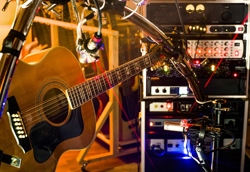 An acoustic guitar in front of a rack of outboard gear, with condenser microphones set up at the soundhole and along the neck.