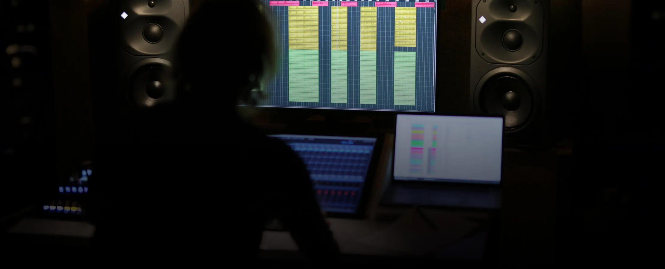 Silhouette of an engineer at a control-room desk, a session open on the screen between two studio monitors.