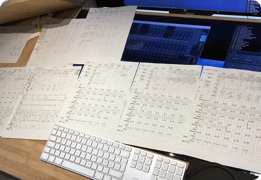 Handwritten mapping and preset sheets spread across a desk in front of a screen showing a sampler instrument.